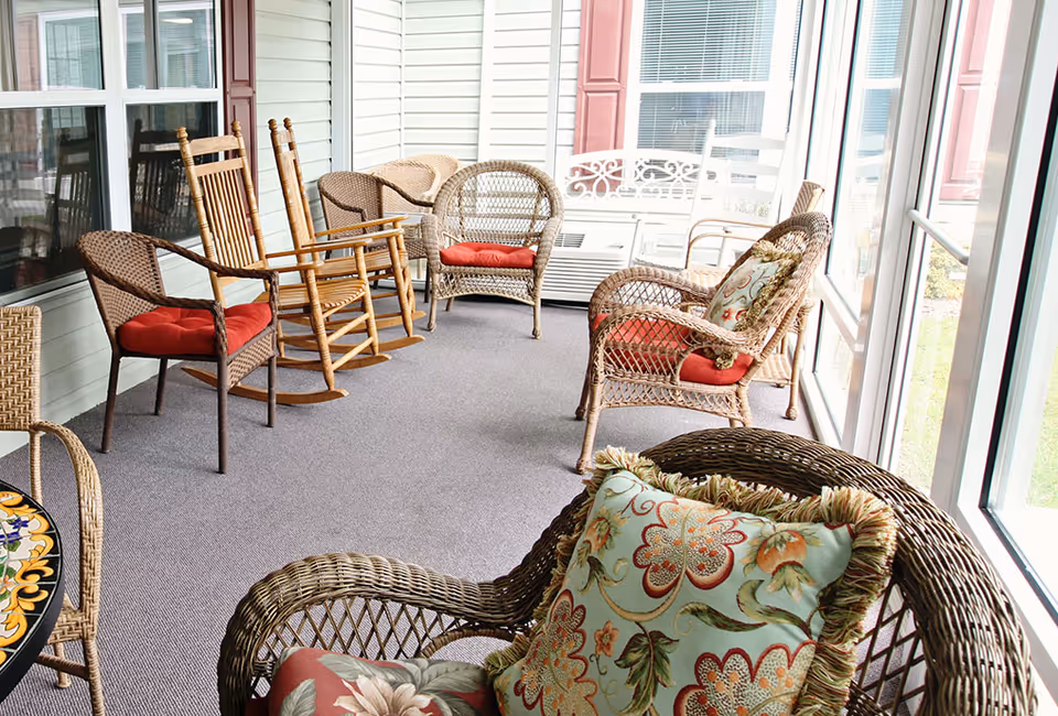 A sunroom or enclosed porch with wicker and wooden rocking chairs arranged along the walls. The chairs have red cushions and some have decorative floral pillows. Large windows let in natural light and offer a view of the outdoors.