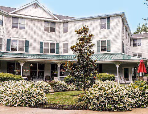Exterior view of a three-story assisted living facility building with white siding and green shutters. The building has a covered porch with columns and hanging flower baskets. In front of the building, there is a landscaped garden with bushes, a small tree, and a red patio umbrella on the right side.