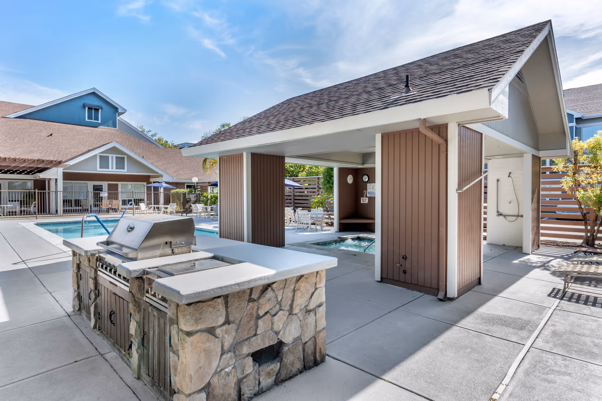Outdoor area of Brookdale Paulin Creek featuring a stone and concrete barbecue grill, a covered hot tub area with clocks on the wall, a shower station, and a swimming pool surrounded by patio furniture and umbrellas under a clear blue sky.