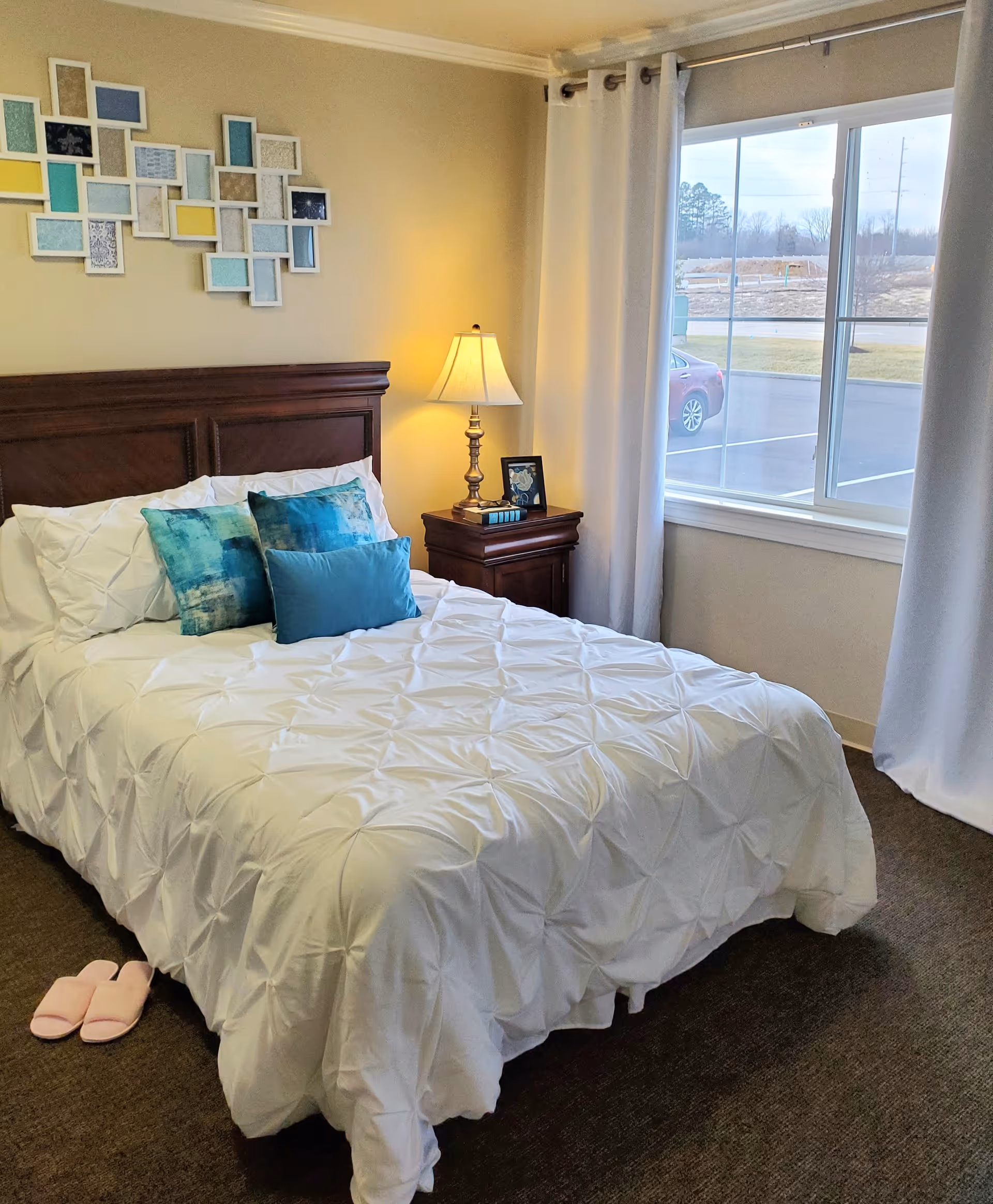 A cozy bedroom with a neatly made bed featuring a white textured comforter and three blue decorative pillows. There is a wooden headboard and a matching wooden nightstand with a lamp, a framed photo, and a small stack of books. A pair of pink slippers is placed on the carpeted floor beside the bed. The room has a large window with white curtains, showing a view of a parking lot and some trees outside. A colorful, abstract wall art piece is hung above the headboard.