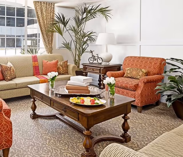 A cozy senior living room with a beige patterned sofa, an orange patterned armchair, a wooden coffee table with books and a plate of fruit, a side table with a lamp and decorative bicycle, and several green plants. The room has large windows with patterned curtains and a carpeted floor with a subtle design.