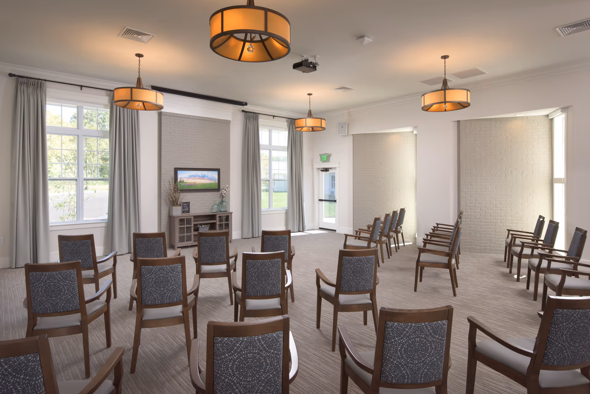 A spacious, well-lit room with multiple rows of wooden chairs arranged facing a small cabinet with decorative items and a wall-mounted TV. The room has large windows with gray curtains, beige walls, and three modern ceiling lights. There is a door at the back leading outside.