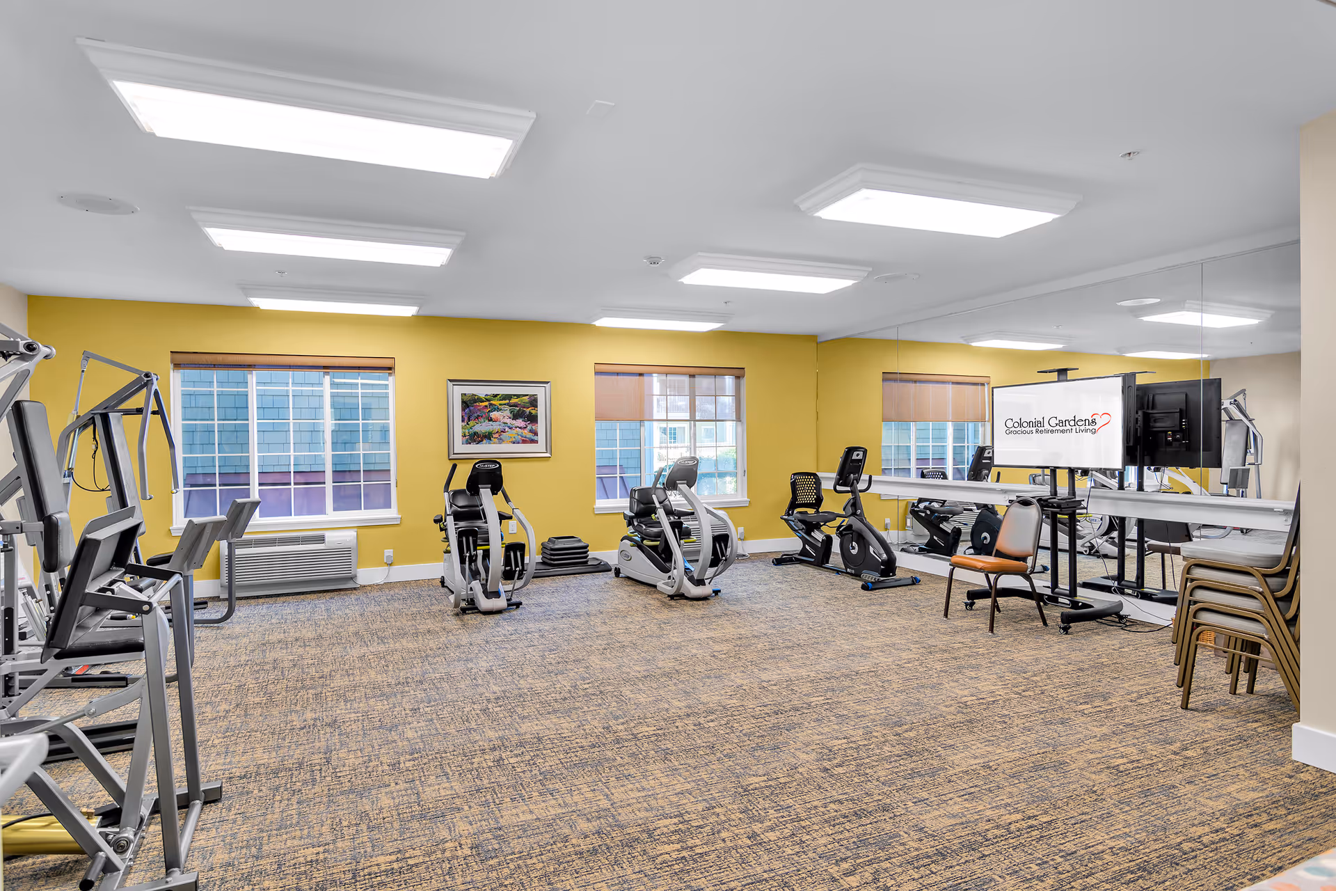 A fitness room with exercise equipment including stationary bikes and weight machines. The room has yellow walls, large windows with blinds, a carpeted floor, and a large mirror on one wall. A TV screen displays the Colonial Gardens Gracious Retirement Living logo. Several stacked chairs and a single chair are also visible.