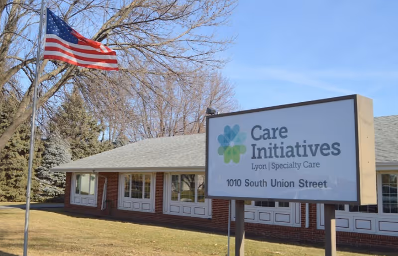 Exterior view of a single-story brick building with multiple windows and a gray roof. In front of the building is a sign that reads 'Care Initiatives Lyon | Specialty Care 1010 South Union Street'. An American flag on a flagpole is also visible on the left side of the image, with leafless trees and a clear blue sky in the background.