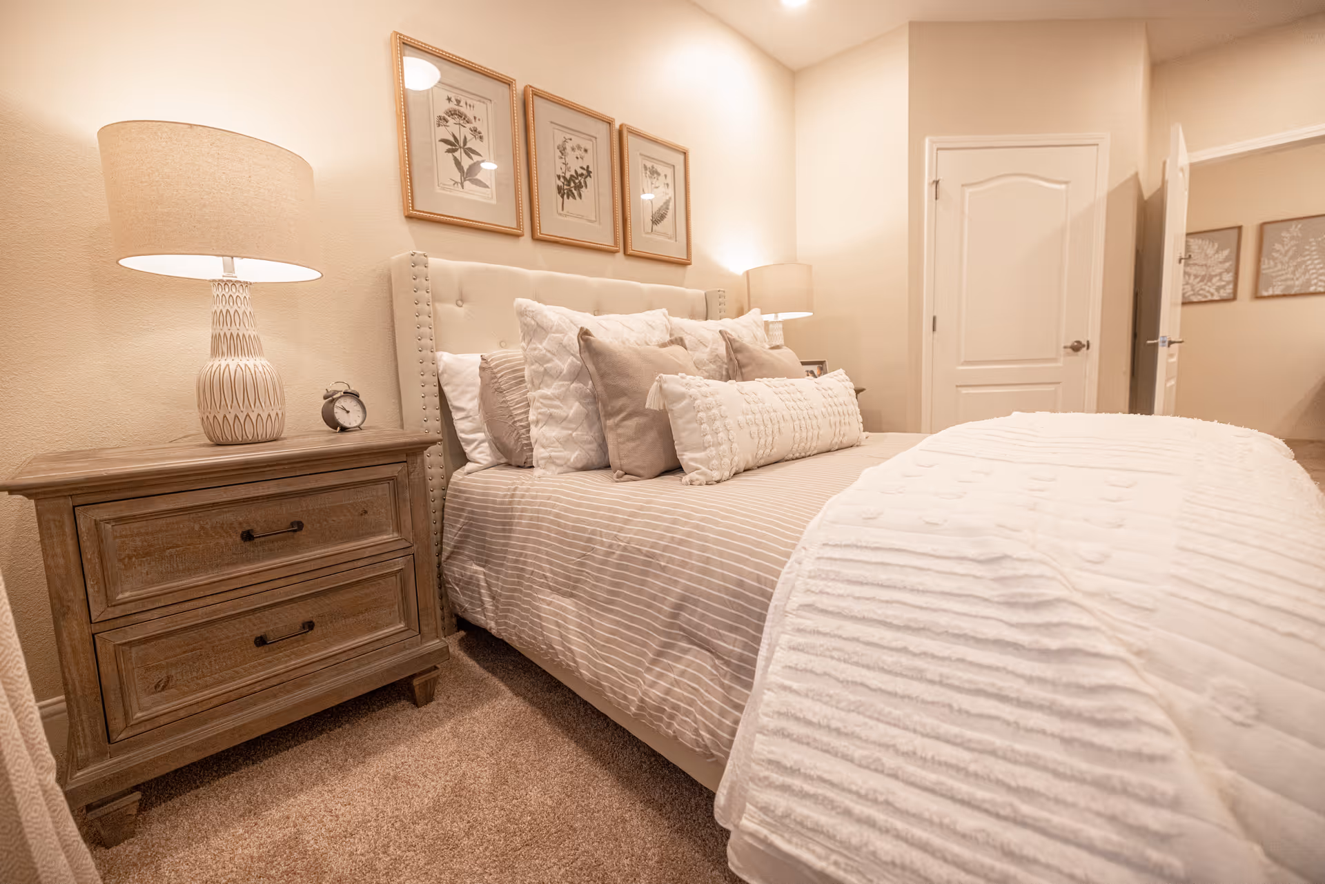 Well-lit bedroom with a tufted headboard bed dressed in layered pillows and a white throw, a wooden nightstand with a lamp and alarm clock, and framed artwork on the wall.