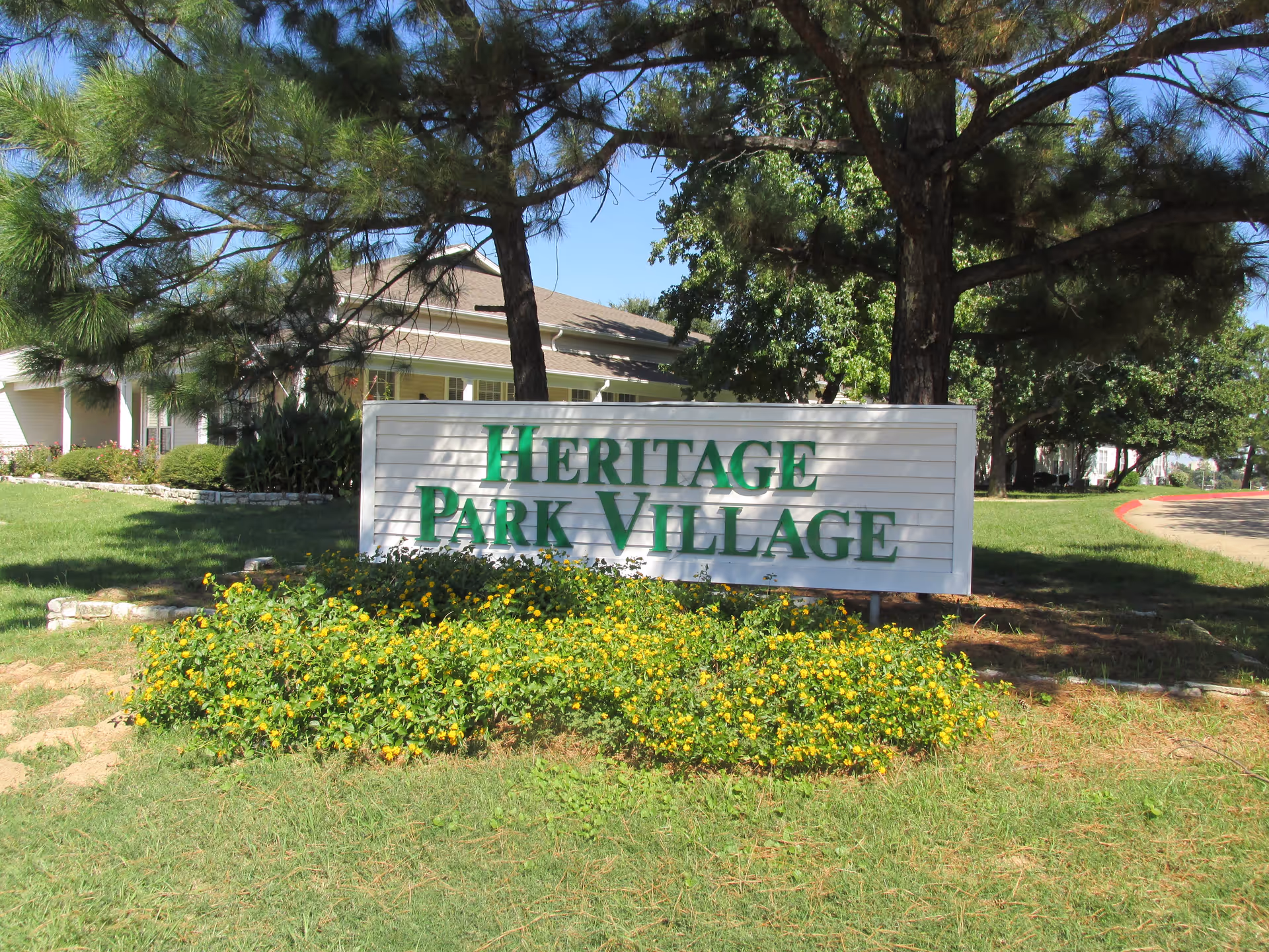 Outdoor view of a sign reading 'Heritage Park Village' surrounded by green grass, yellow flowers, and trees with a building partially visible in the background under a clear blue sky.