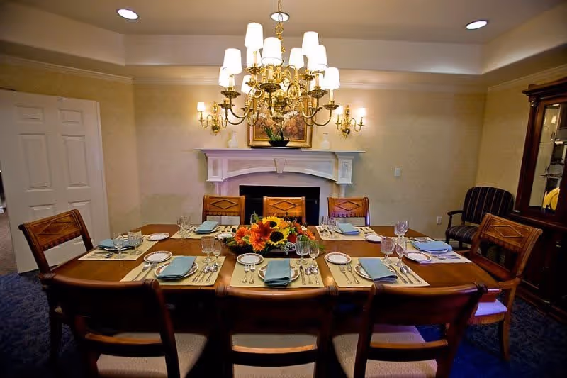A formal dining room with a wooden table set for eight people, featuring plates, glasses, silverware, and blue napkins. A floral centerpiece with sunflowers decorates the middle of the table. The room has a fireplace with a white mantel, wall sconces with lights, a chandelier above the table, and a wooden cabinet on the right side.
