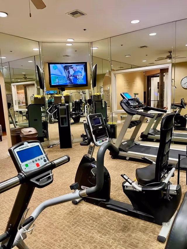 Interior fitness room with recumbent exercise bikes, treadmills, mirrored walls and a wall-mounted TV.