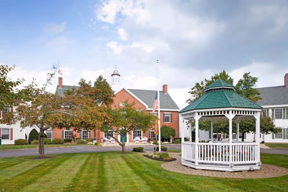 Outdoor view of a senior living facility with a white gazebo featuring a green roof in the foreground, well-maintained green lawn, trees, and a brick building with white trim and an American flag at half-mast in the background under a partly cloudy sky.
