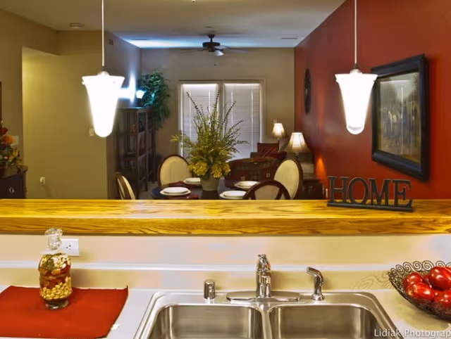 View from a kitchen sink area looking into a dining and living room space. The kitchen counter has a glass jar with nuts, a red placemat, and a bowl of red apples. The dining table is set with plates and has a floral centerpiece. The living room has a ceiling fan, lamps, and a red accent wall with a framed picture and a decorative 'HOME' sign.