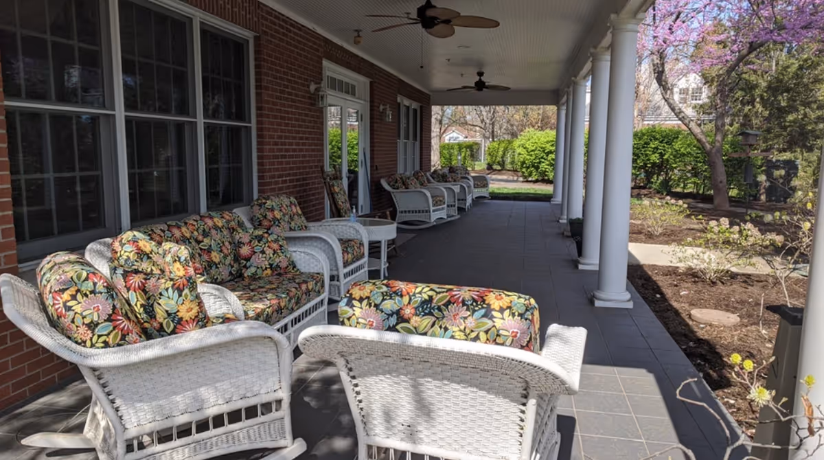 Covered outdoor patio with white wicker chairs with floral cushions along a brick building overlooking a garden.