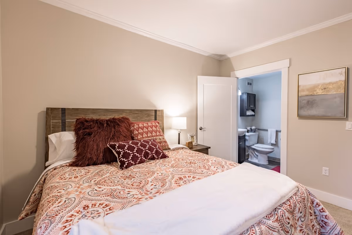 A furnished bedroom with a patterned bed, decorative pillows, a bedside lamp, and an open door revealing an attached bathroom.