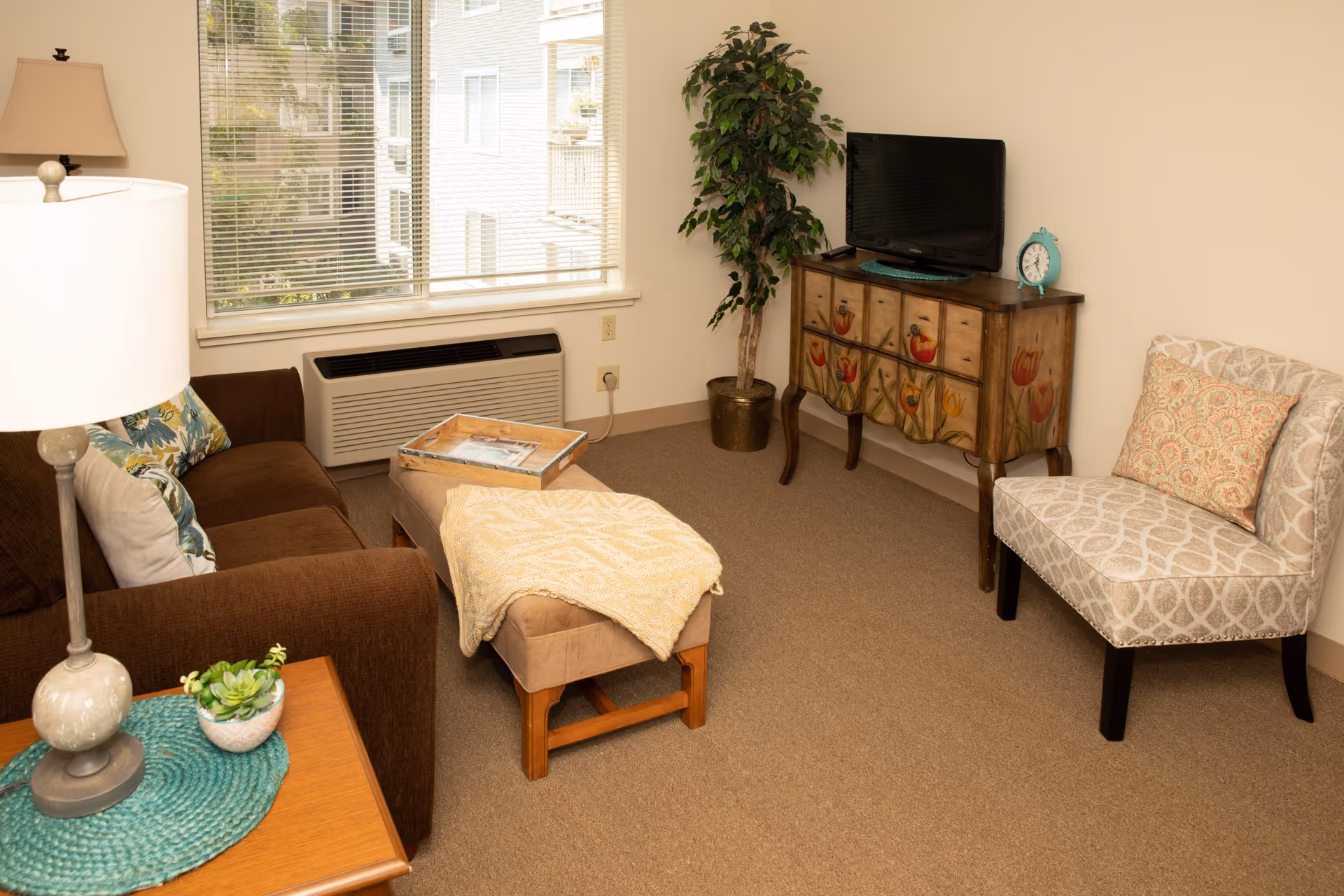 Cozy living room with a brown sofa, ottoman, patterned accent chair, small TV on a decorative cabinet, and a window with blinds.