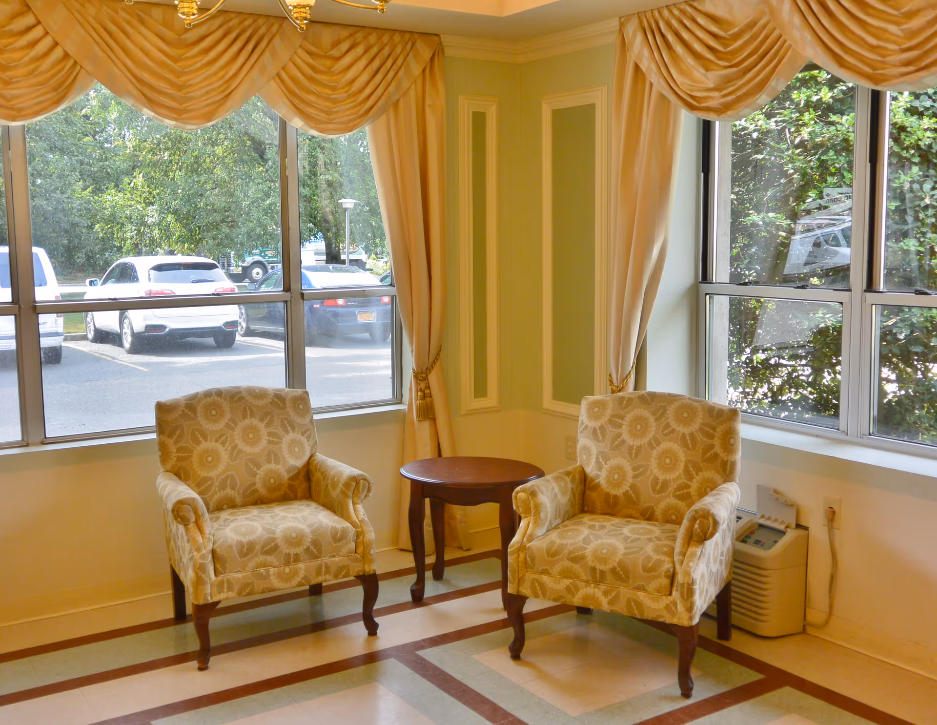 A cozy sitting area with two patterned armchairs and a small round wooden table between them, positioned near large windows with beige curtains and valances. Outside the windows, several parked cars and greenery are visible.