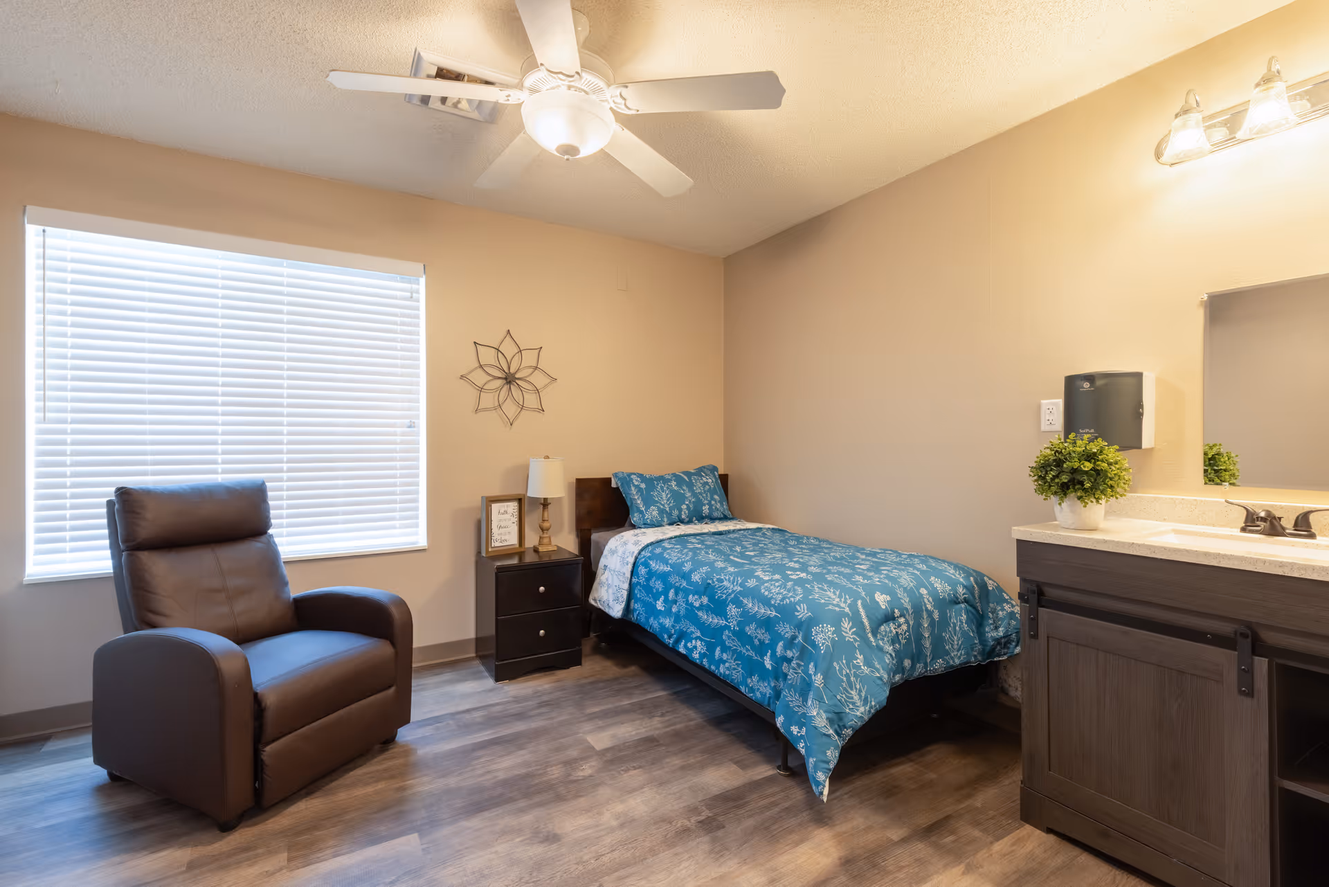 A cozy bedroom with a single bed covered in a blue floral comforter, a dark brown recliner chair near a window with white blinds, a small nightstand with a lamp and framed picture, a ceiling fan, and a sink with a mirror and potted plant on the right side.