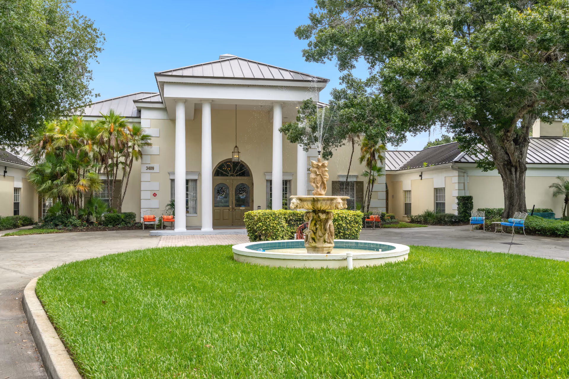 Front exterior view of Summerfield Senior Living building with a circular driveway, green lawn, a decorative fountain in the center, and trees and shrubs surrounding the entrance.