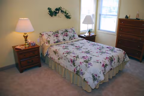 A bedroom with a bed covered in floral bedding, two wooden nightstands each with a lamp, a wooden dresser, and a window letting in natural light. The walls are painted light yellow and there is a decorative plant hanging above the bed.