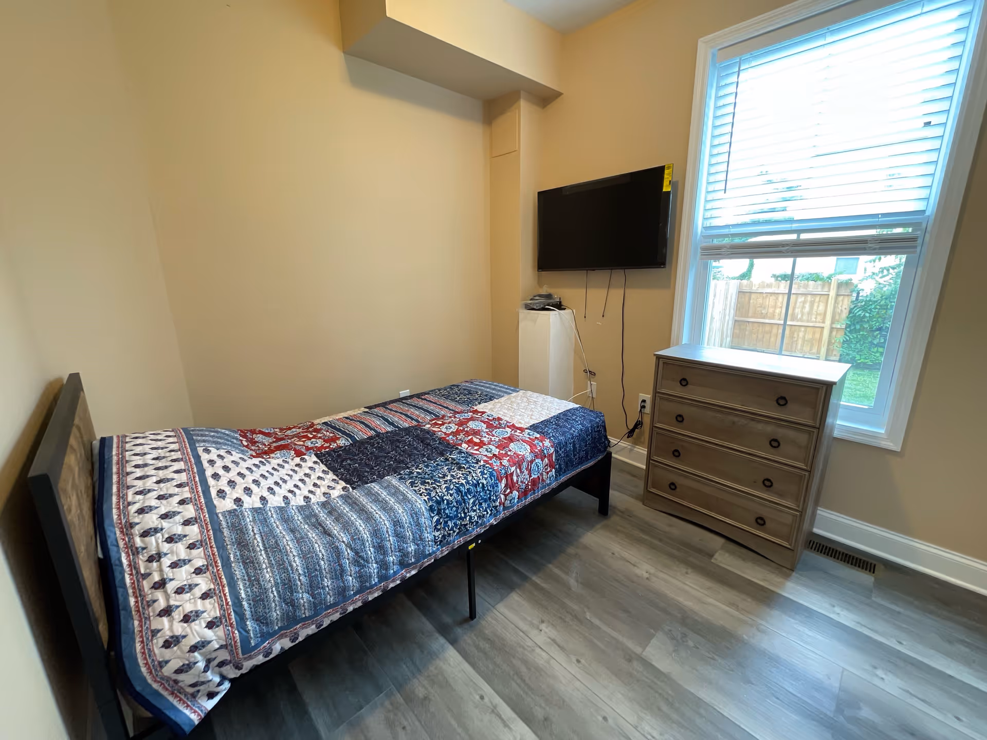 A small bedroom with a single bed covered with a patchwork quilt in blue, red, and white colors. There is a wooden dresser with four drawers next to a window with white blinds, and a flat-screen TV mounted on the wall above a small white pedestal. The room has light beige walls and wood-look flooring.