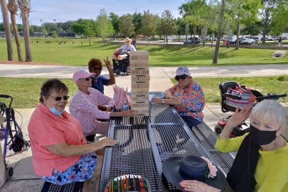 A group of elderly women sitting around a metal picnic table outdoors in a park-like setting. They are playing a game with large wooden blocks stacked in a tower. Some women are wearing hats and sunglasses, and one woman is waving at the camera. There are trees, grass, and a sidewalk in the background, along with a person in a wheelchair and parked cars.