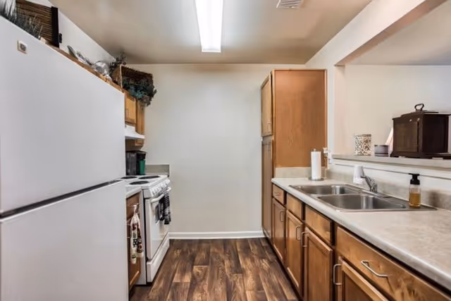 Galley-style kitchen with wooden cabinets, white appliances, a double sink and wood-look flooring.