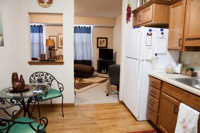 View of a small kitchen area with wooden cabinets, a white refrigerator, and a sink on the right. On the left, there is a round glass table with two green cushioned wrought iron chairs. Through an open doorway, a living room area is visible with a TV on a stand, a brown ottoman, and blue curtains on the windows.