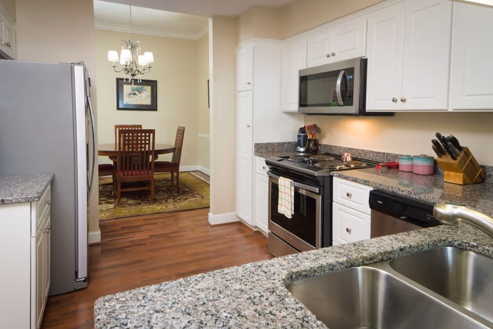 A modern kitchen with granite countertops, white cabinets, a stainless steel double sink, stove, microwave, dishwasher, and refrigerator. In the background, there is a dining area with a round wooden table, four chairs, a chandelier, and a framed picture on the wall.