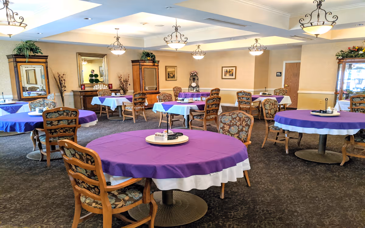 A dining room with multiple round and square tables covered with white and purple tablecloths. Each table is surrounded by wooden chairs with floral upholstery. The room has beige walls, carpeted floors, decorative plants, framed pictures, and several hanging light fixtures. There is a mirror and wooden cabinets along the walls.