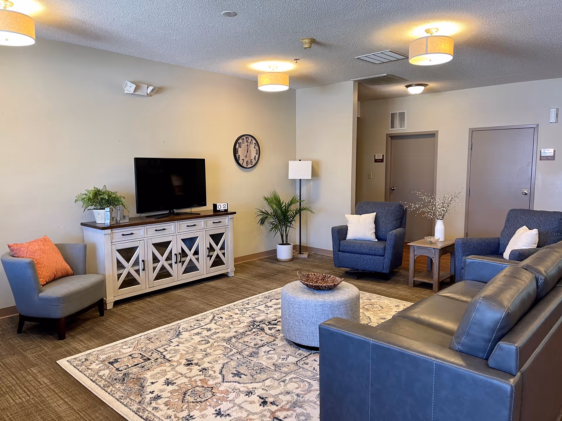 A cozy living room area in Limestone Lodge featuring a gray leather sofa, two blue armchairs with white pillows, a gray armchair with an orange pillow, a patterned area rug, and a round gray ottoman. A white wooden TV stand holds a flat-screen television, a plant, and a small calendar. The walls are light beige with a round clock and a floor lamp next to a potted plant. Two closed doors are visible in the background under ceiling lights.