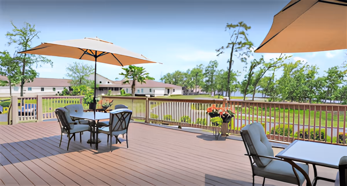Outdoor patio area with tables and chairs under large beige umbrellas, overlooking a green lawn with trees and buildings in the background under a clear blue sky.