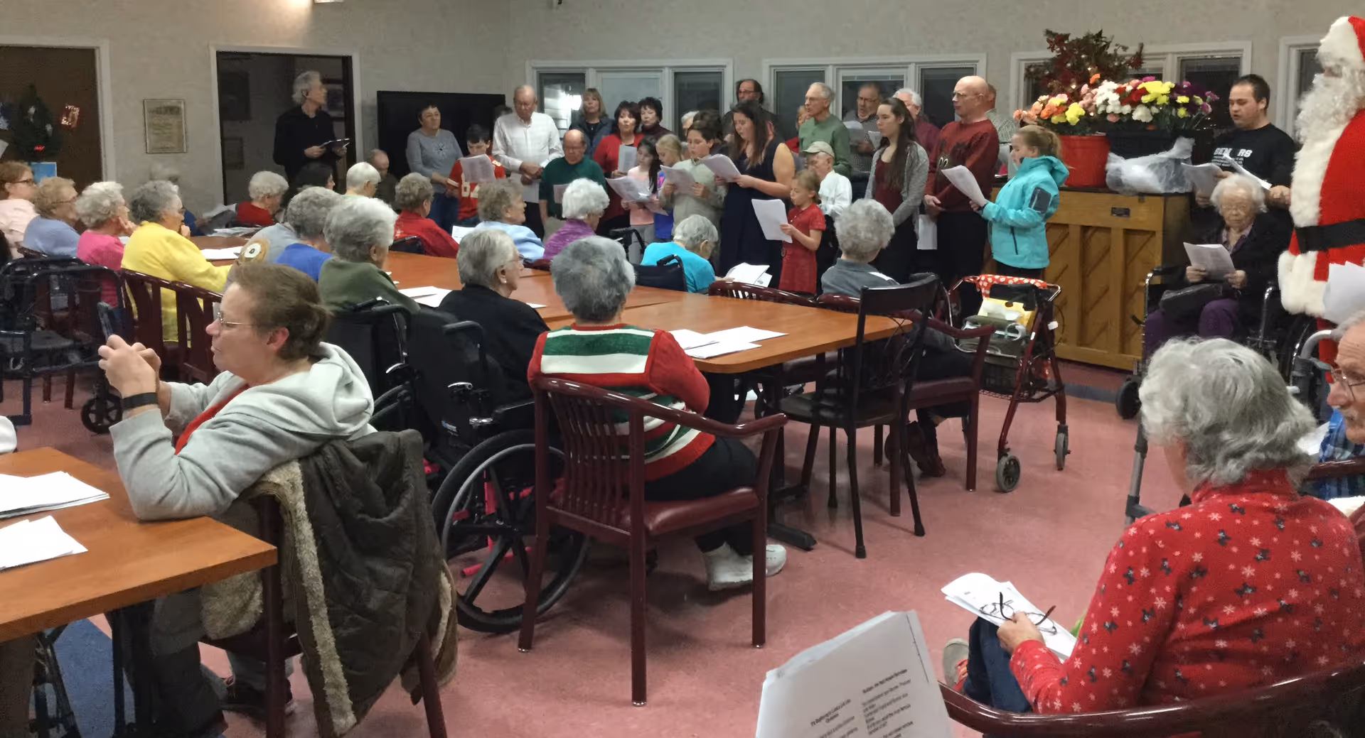 Senior residents seated around tables in a community room watching a choir perform.