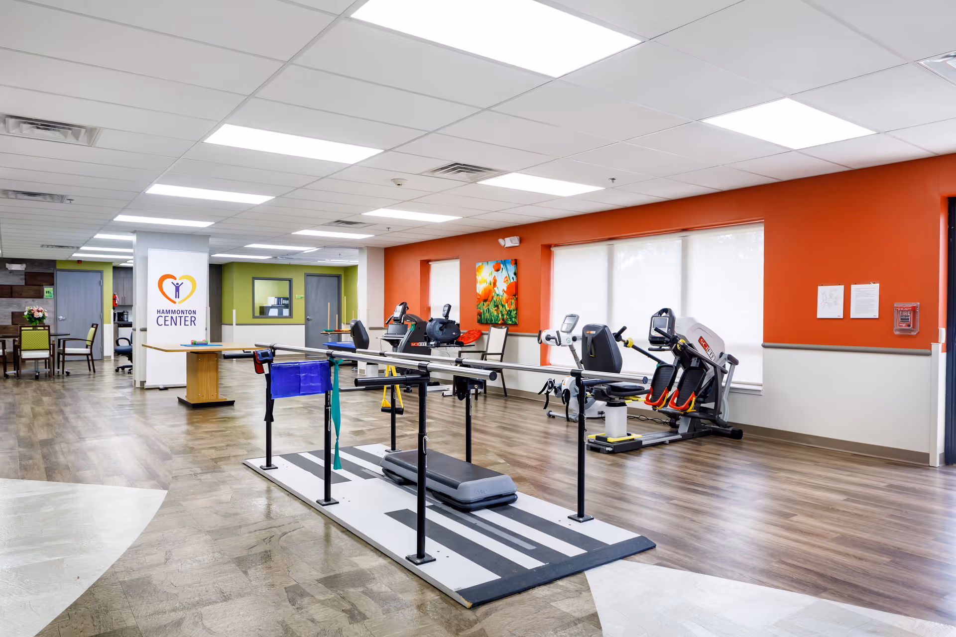 A spacious rehabilitation or exercise room in Hammonton Center featuring parallel bars for walking exercises, several stationary exercise bikes, a colorful painting on an orange accent wall, and a seating area with chairs and tables in the background.