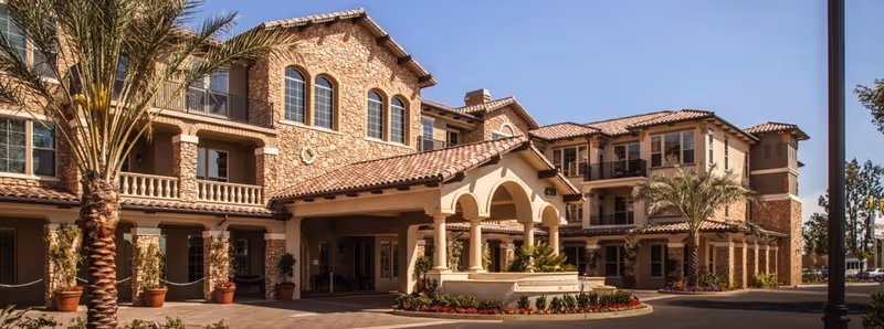 Exterior view of a large, multi-story senior living facility with stone and stucco facade, arched windows, and a covered entrance. Palm trees and potted plants decorate the front area under a clear blue sky.