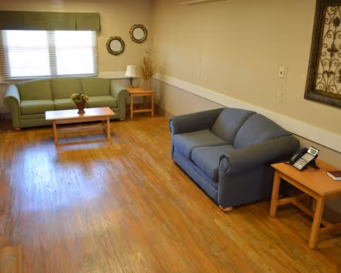 A cozy living room with wooden flooring featuring two sofas, one green and one blue, a wooden coffee table with a flower vase, two side tables with a lamp and a telephone, a window with blinds, and decorative wall hangings.