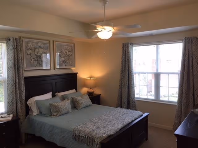 A cozy bedroom with a dark wooden bed frame, light blue bedding, and multiple pillows. There are two framed floral artworks above the bed, a ceiling fan with a light, a bedside table with a lamp, and two windows with patterned curtains allowing natural light into the room.