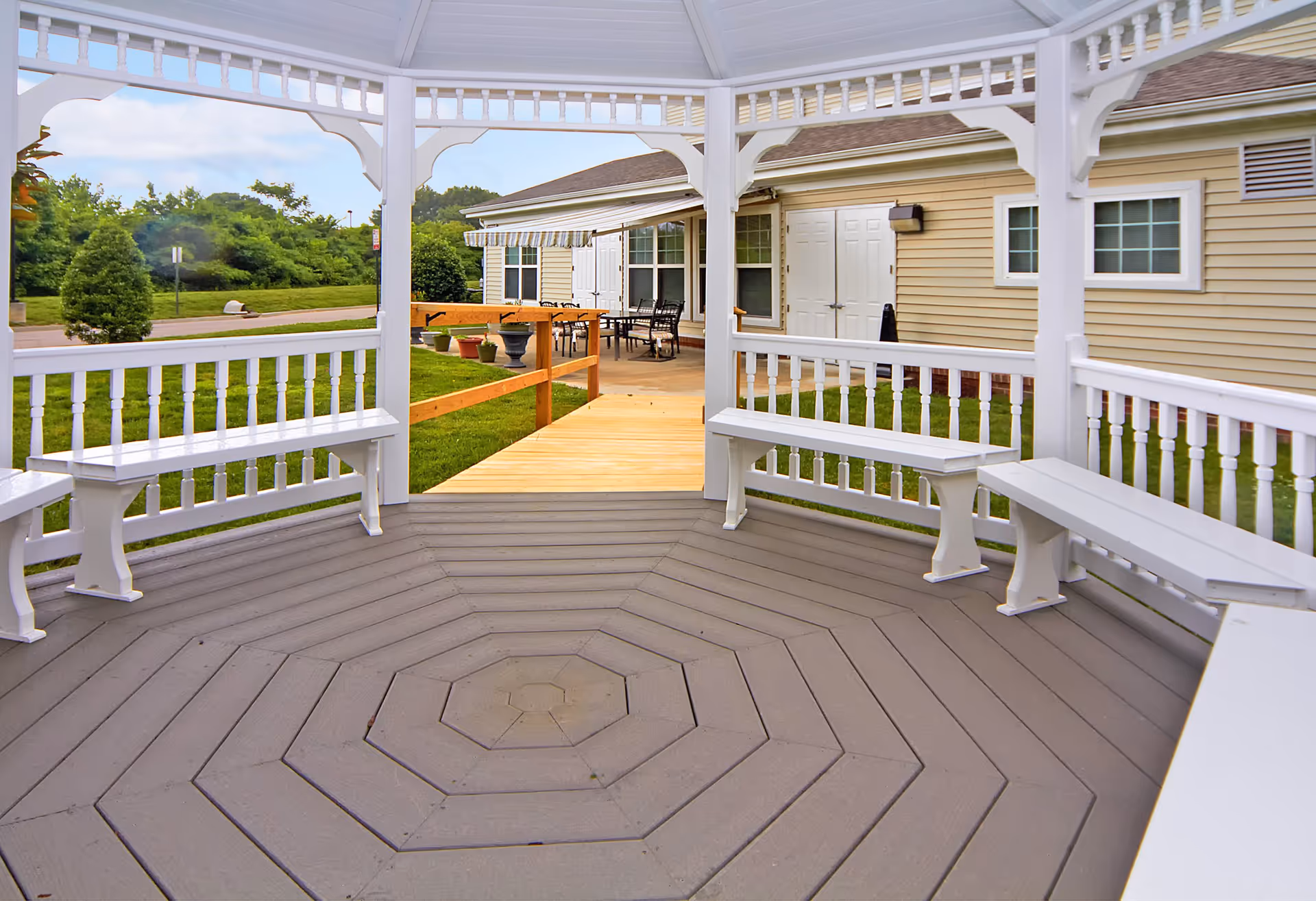 View from inside a white wooden gazebo with built-in benches, looking out towards a beige building with white doors and windows. There is a wooden ramp leading from the gazebo to a patio area with outdoor tables and chairs. Green grass and trees are visible in the background under a partly cloudy sky.