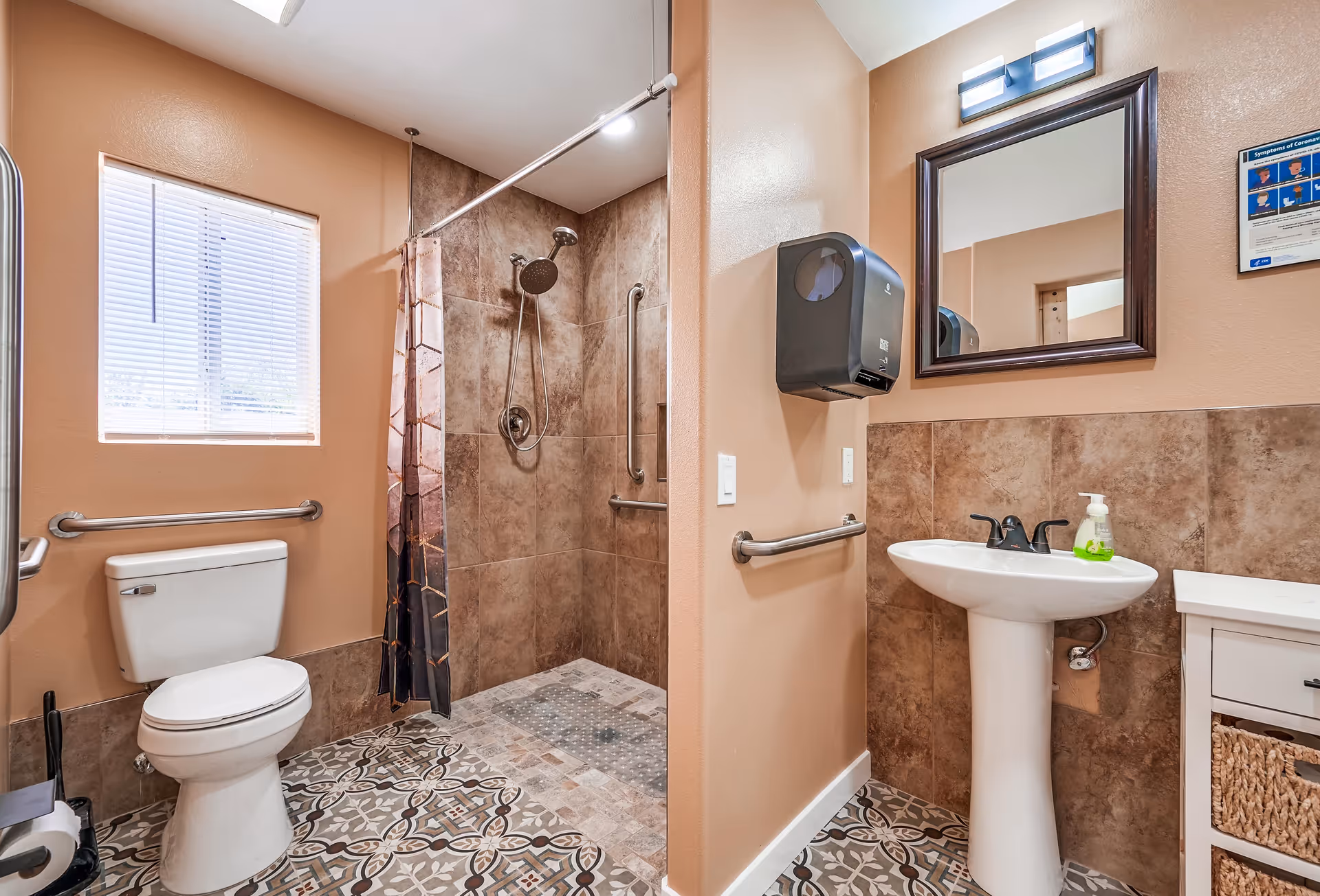 A clean and accessible bathroom with beige walls and patterned tile flooring. The bathroom features a white toilet with a grab bar beside it, a walk-in shower with a handheld showerhead and grab bars, a white pedestal sink with a soap dispenser, a mirror above the sink, and a black paper towel dispenser mounted on the wall. There is a window with blinds allowing natural light into the room.