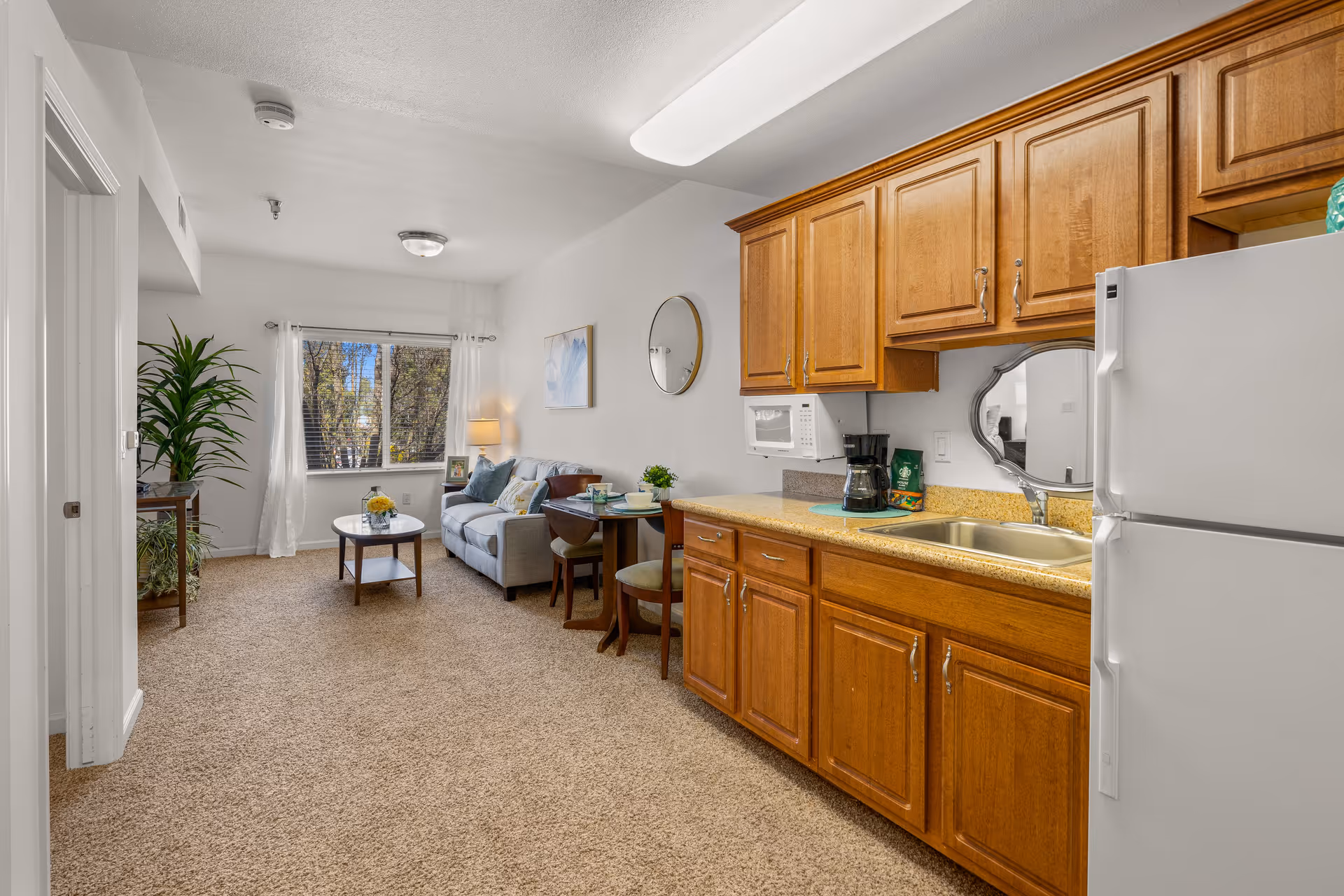 Interior view of a senior living facility apartment featuring a small kitchen area with wooden cabinets, a white refrigerator, a microwave, and a coffee maker. Adjacent to the kitchen is a cozy living room with a gray sofa, a round wooden coffee table, a small dining table with two chairs, a floor lamp, and a large window with white curtains letting in natural light. The room has beige carpet flooring and white walls decorated with a round mirror and a framed picture.