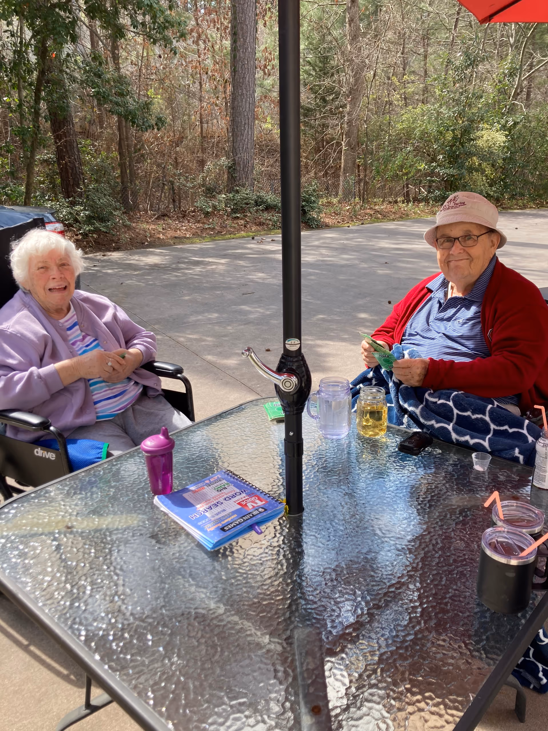 Two elderly individuals sitting outside at a glass table with a large umbrella pole in the center. One person is in a wheelchair wearing a light purple jacket and striped shirt, smiling. The other person is wearing a red jacket, blue shirt, glasses, and a light-colored hat, also smiling. On the table are various items including a purple sippy cup, a book, a jar with a yellow drink, a water pitcher, and two cups with straws. The background shows a wooded area with trees and a paved surface.
