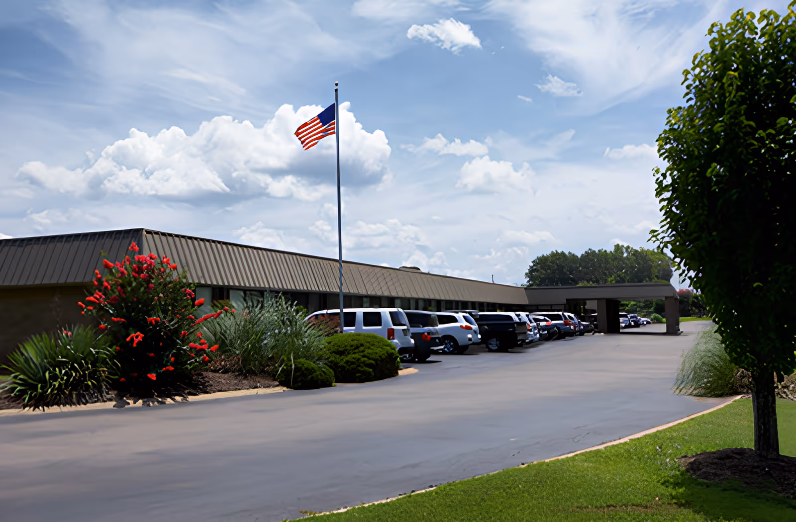Exterior view of a single-story building with a brown roof and multiple parked cars along the driveway. An American flag is flying on a flagpole near the building, with green shrubs and red flowers in the landscaped area. The sky is partly cloudy with blue patches.