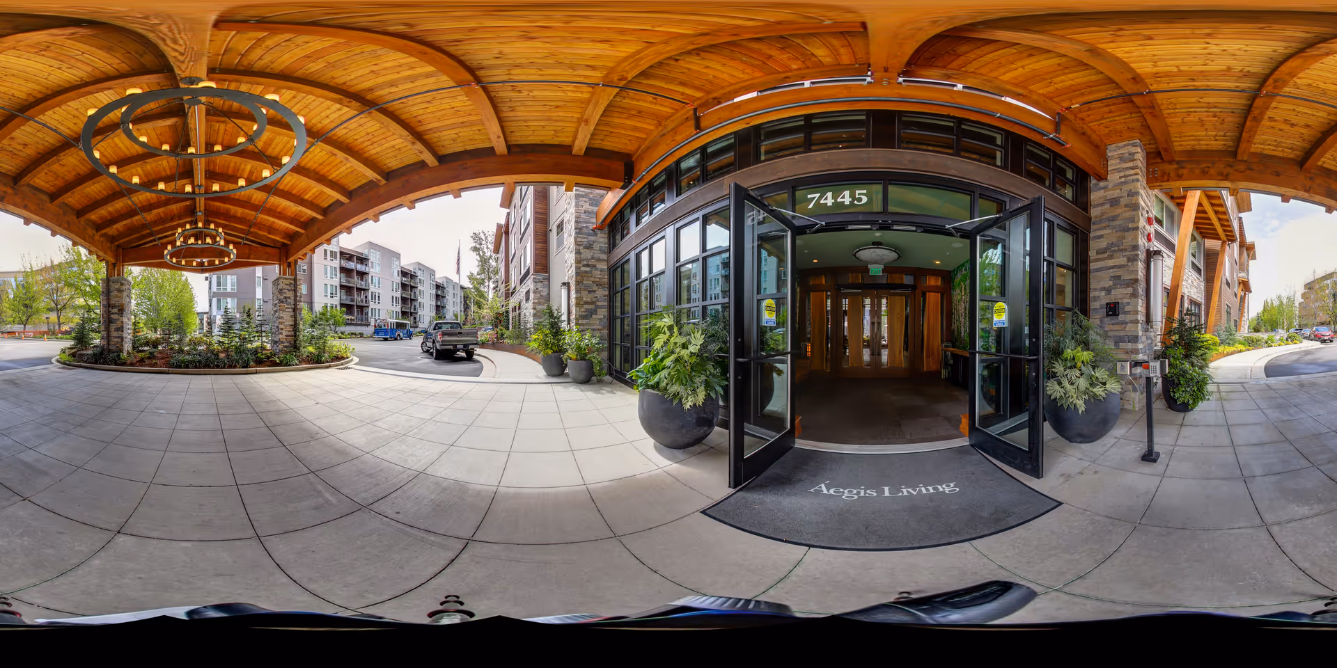 Covered entrance area of Aegis Living Mercer Island facility with wooden ceiling and circular light fixtures. Glass double doors open into the building, with potted plants on either side and a welcome mat that reads 'Aegis Living'. Surrounding the entrance are landscaped areas and multi-story residential buildings in the background.