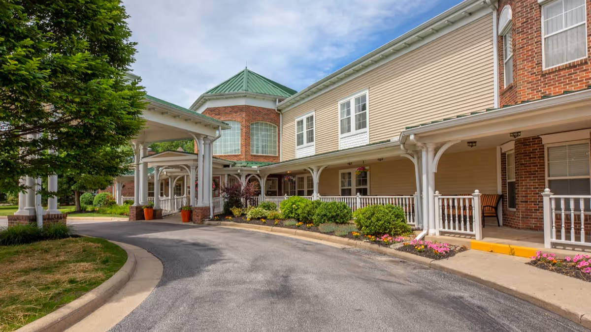 Exterior view of a senior living facility with a covered entrance, beige siding, brick accents, white railings, and a green roof. There are landscaped bushes and flowers along the sidewalk and a tree on the left side.