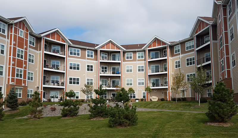 Exterior view of a multi-story senior living community building with balconies, surrounded by a well-maintained lawn and small trees under a partly cloudy sky.