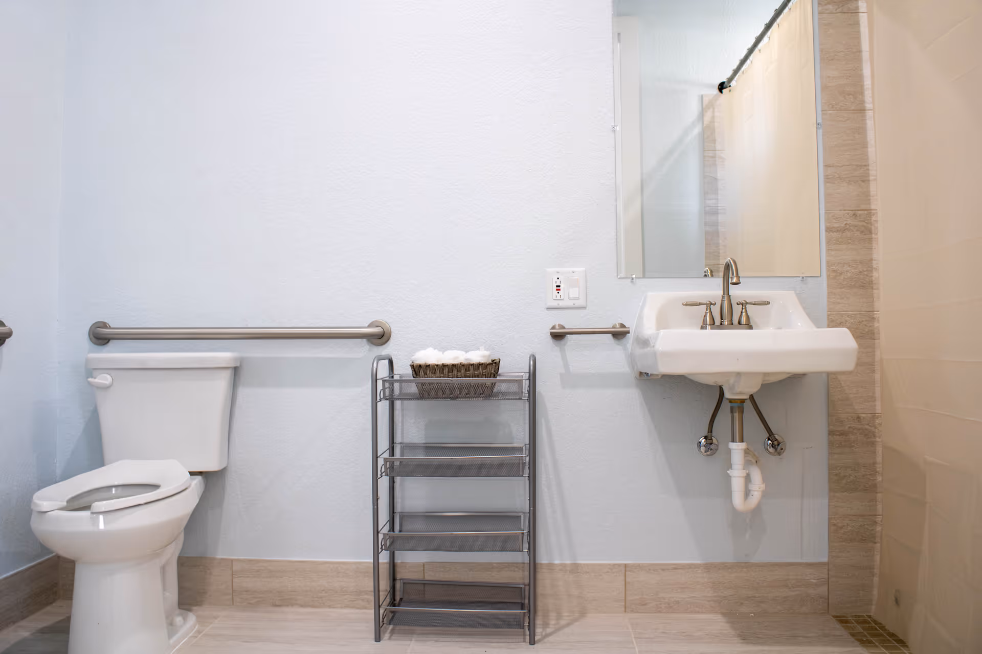A clean and accessible bathroom featuring a white toilet with a safety grab bar on the wall behind it, a metal shelving unit with rolled white towels, a wall-mounted sink with a faucet and a mirror above it, and a shower area with a beige curtain.