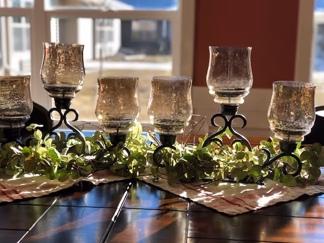 A decorative centerpiece on a wooden table featuring six glass candle holders with black metal stands, surrounded by green leafy garland and placed on a beige and red checkered cloth. Sunlight streams through a window in the background.