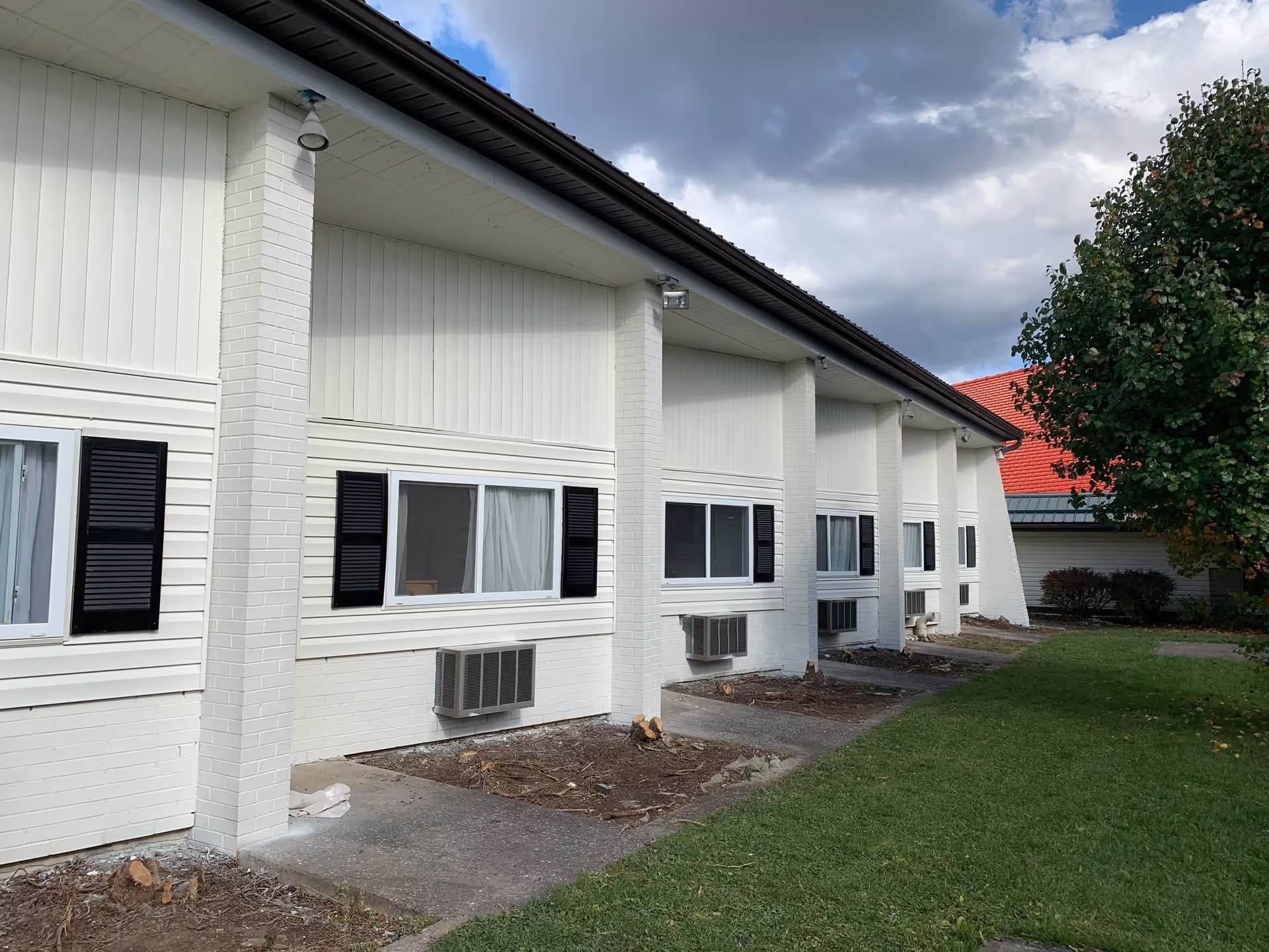 Exterior side view of a white senior living facility building with multiple windows, each having black shutters and air conditioning units below. There is a green lawn and a tree on the right side under a partly cloudy sky.