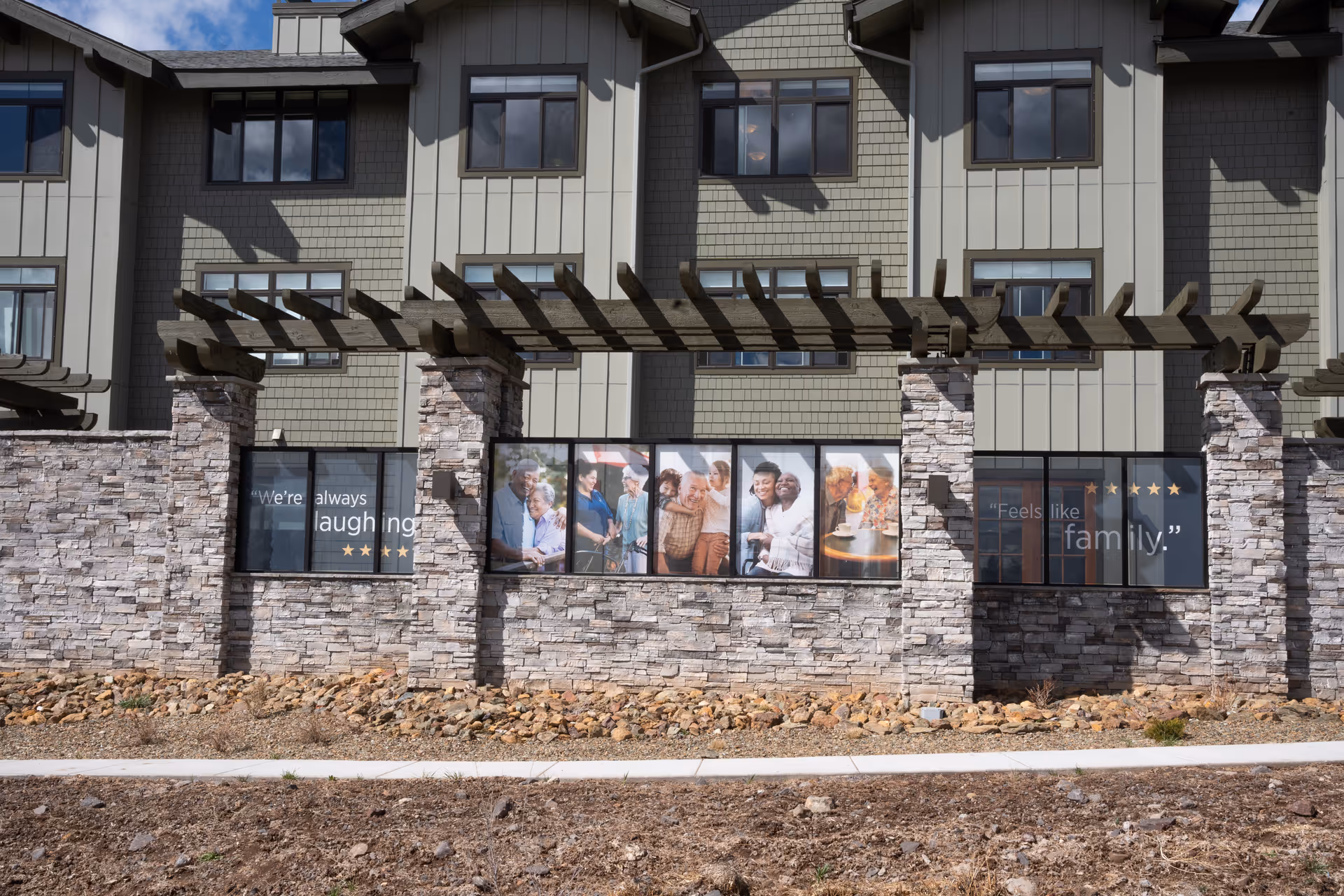 Stone-faced exterior of a senior living building with a wooden pergola and windows displaying photos and quotes.