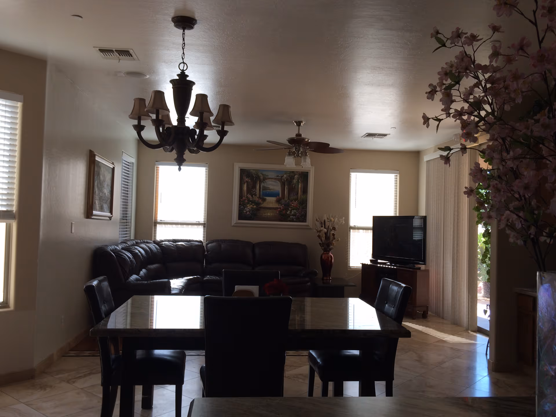 Interior view of a living room and dining area with a dark leather sectional sofa, a flat-screen TV on a wooden stand, a dining table with four chairs, a chandelier hanging from the ceiling, and large windows with blinds letting in natural light. There is a painting on the wall and a vase with flowers near the TV.
