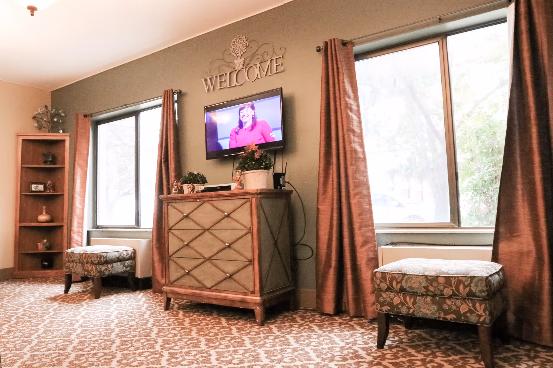 A cozy living room area with two patterned upholstered stools, a wooden cabinet with plants on top, a wall-mounted TV showing a woman in a red top, and a decorative 'WELCOME' sign above the TV. The room has large windows with brown curtains and a patterned carpet.