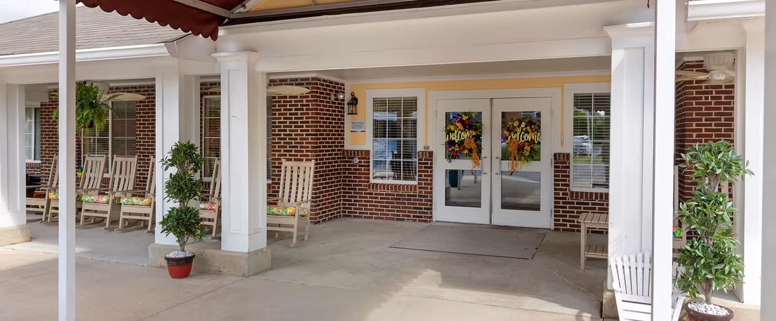 Entrance to a building with double glass doors decorated with colorful welcome signs. The exterior features red brick walls, white columns, and a covered porch area with several wooden rocking chairs and potted plants.