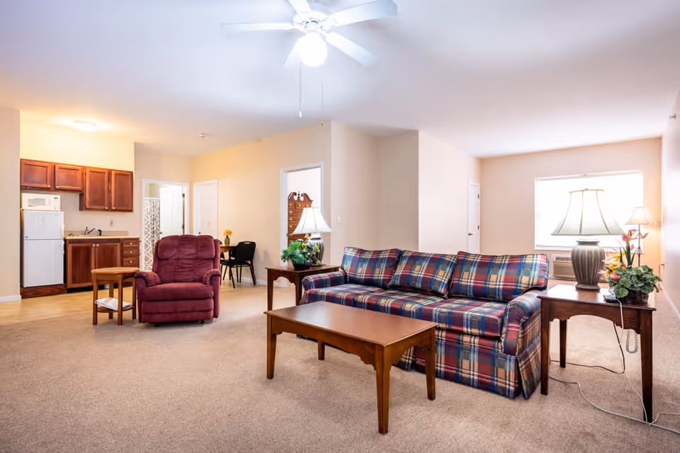Spacious assisted living apartment living room with a plaid sofa, red recliner, coffee table, side tables with lamps, and a small kitchenette in the background.